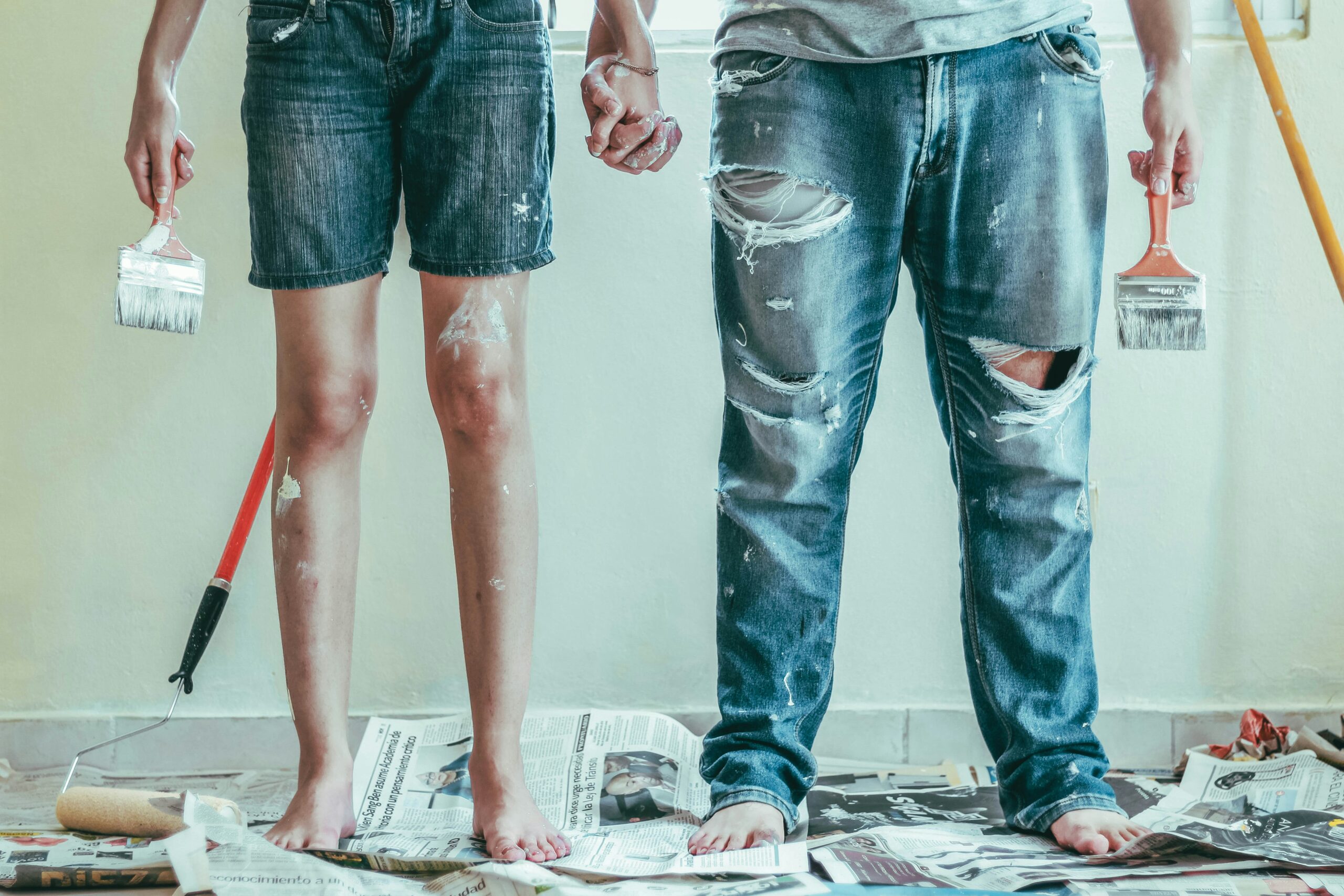 a couple painting a wall, with them holding a paint brush and a paint roller on the floor with a paint tray, showing a DIY home improvement project in progress.